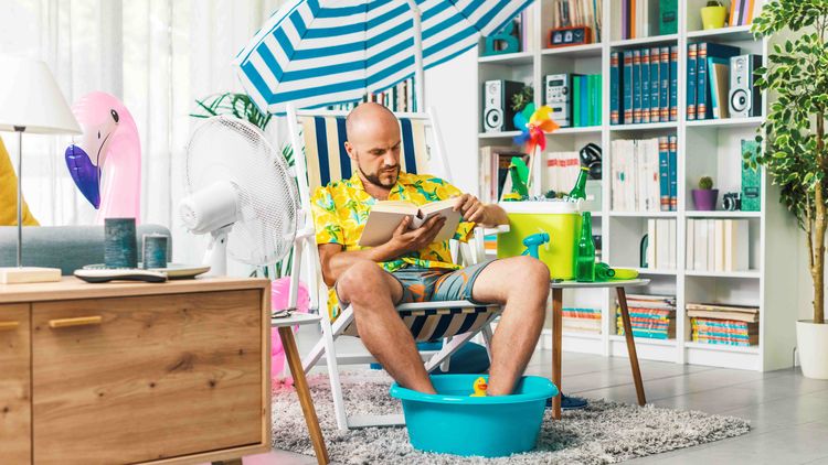 Mann verbringt seine Ferien zu Hause und liest ein Buch, dabei stehen seine Füße in einer Schüssel mit Wasser, neben ihm steht ein Ventilator und er sitzt in der Wohnung auf einem Strandstuhl mit Sonnenschirm.